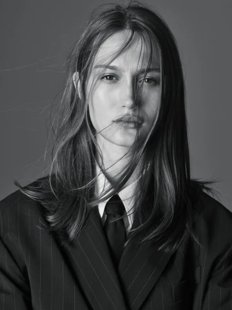Monochrome portrait of a young woman with flowing hair, wearing a striped blazer, white collared shirt, and black tie, gazing at the camera.