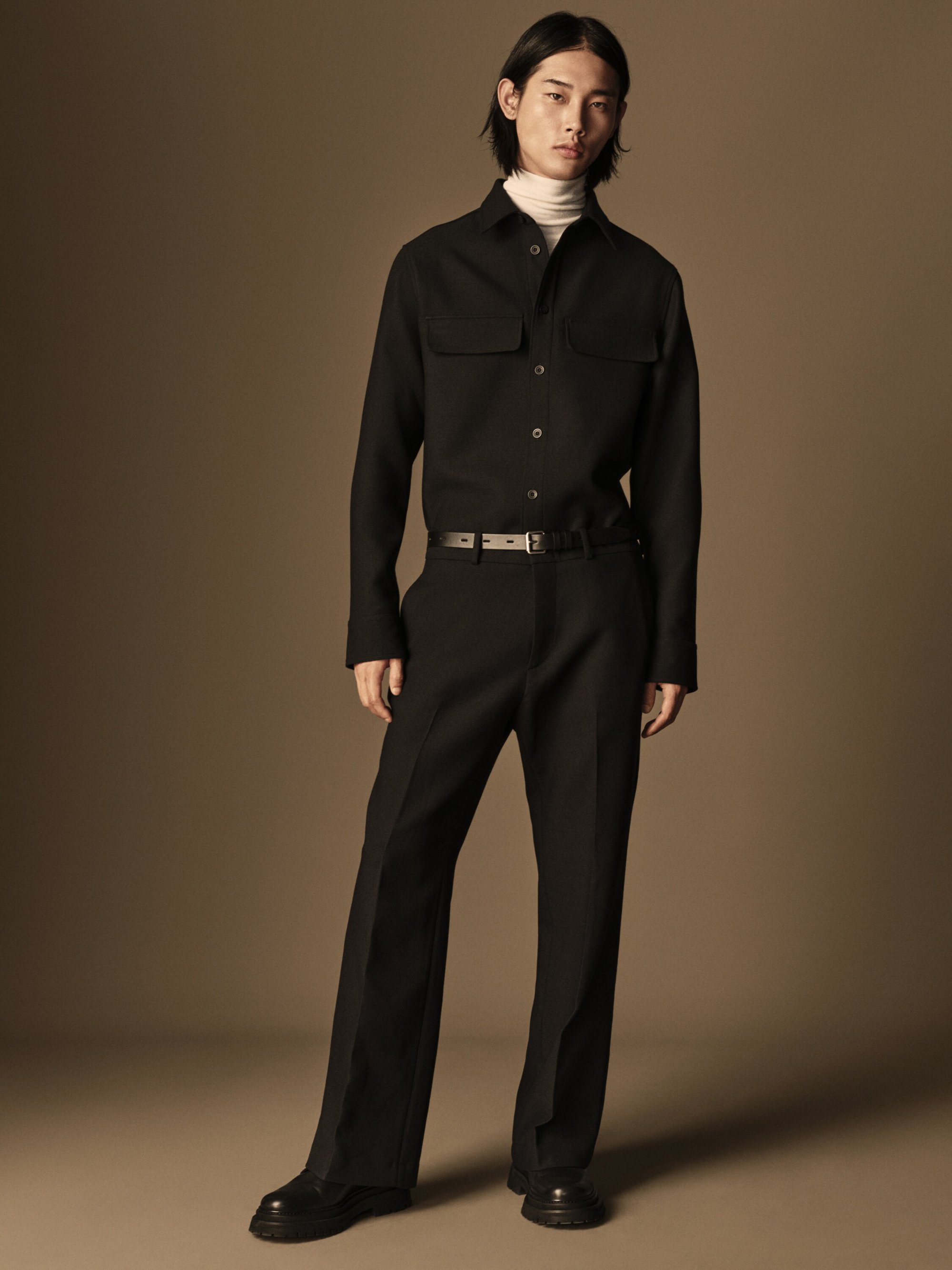 Asian man in a black button-down shirt and black trousers stands in a neutral studio, fashionably posing with a subtle expression.
