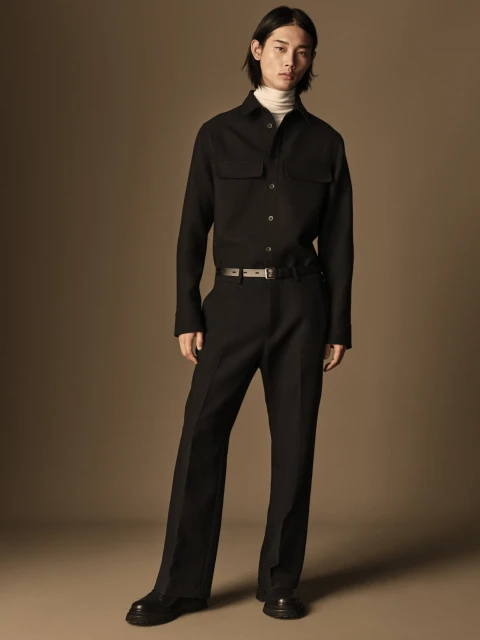 Asian man in a black button-down shirt and black trousers stands in a neutral studio, fashionably posing with a subtle expression.