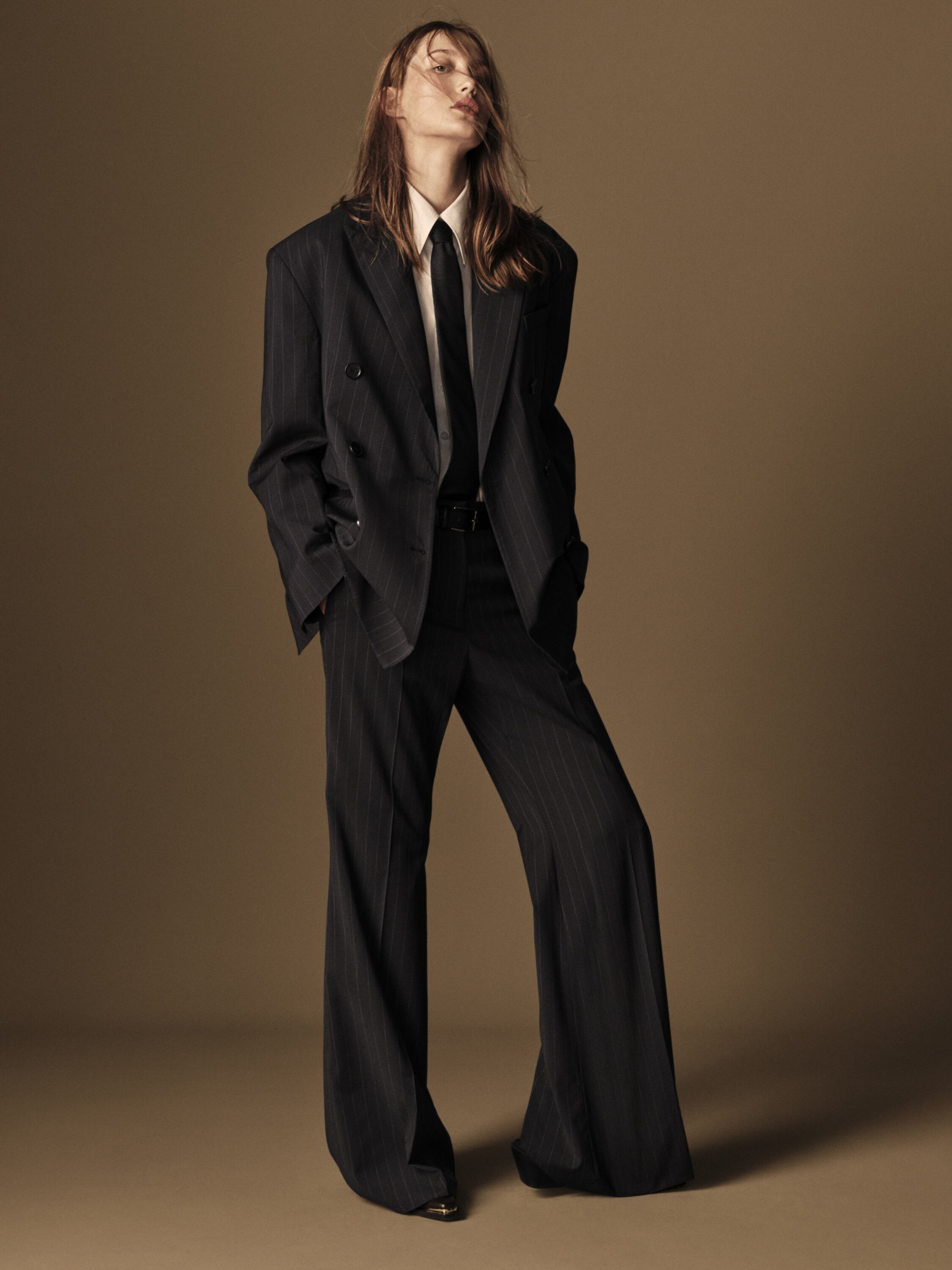 Woman in an oversized pinstripe suit, standing confidently in a studio with warm lighting. Fashion editorial with a vintage-inspired look.