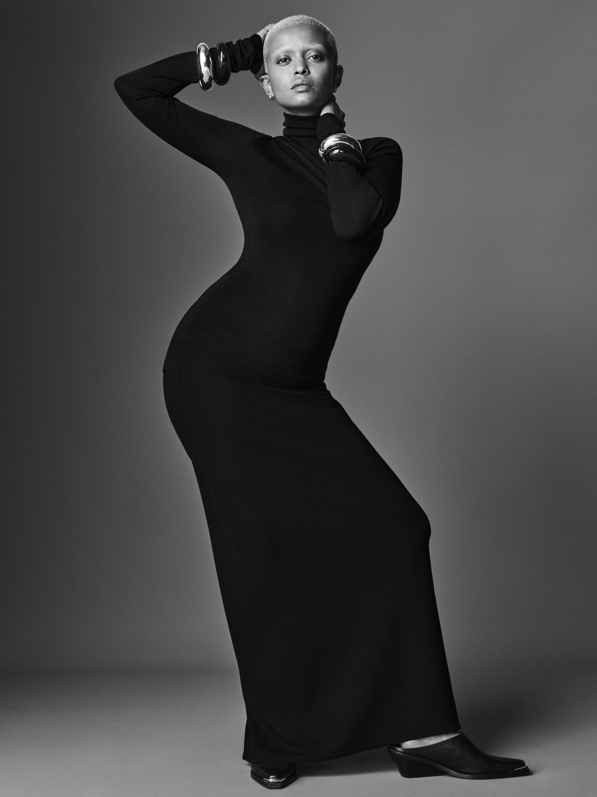 Woman with light hair poses in long black turtleneck dress with silver cuff bracelets in a studio with neutral grey background.
