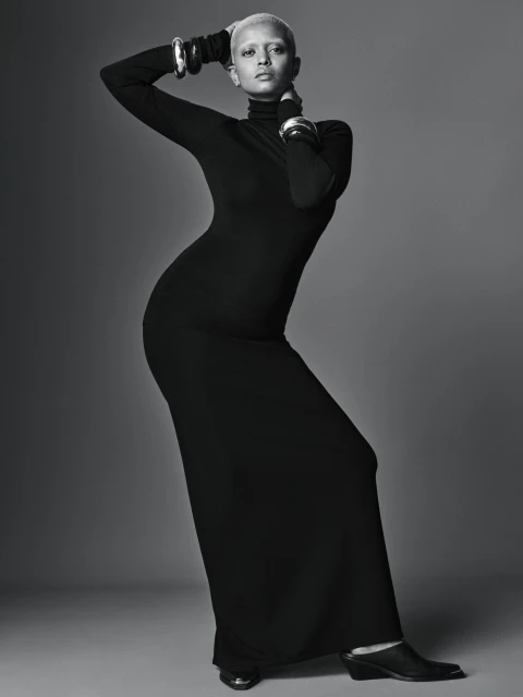 Woman with light hair poses in long black turtleneck dress with silver cuff bracelets in a studio with neutral grey background.