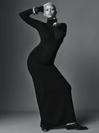 Woman with light hair poses in long black turtleneck dress with silver cuff bracelets in a studio with neutral grey background.
