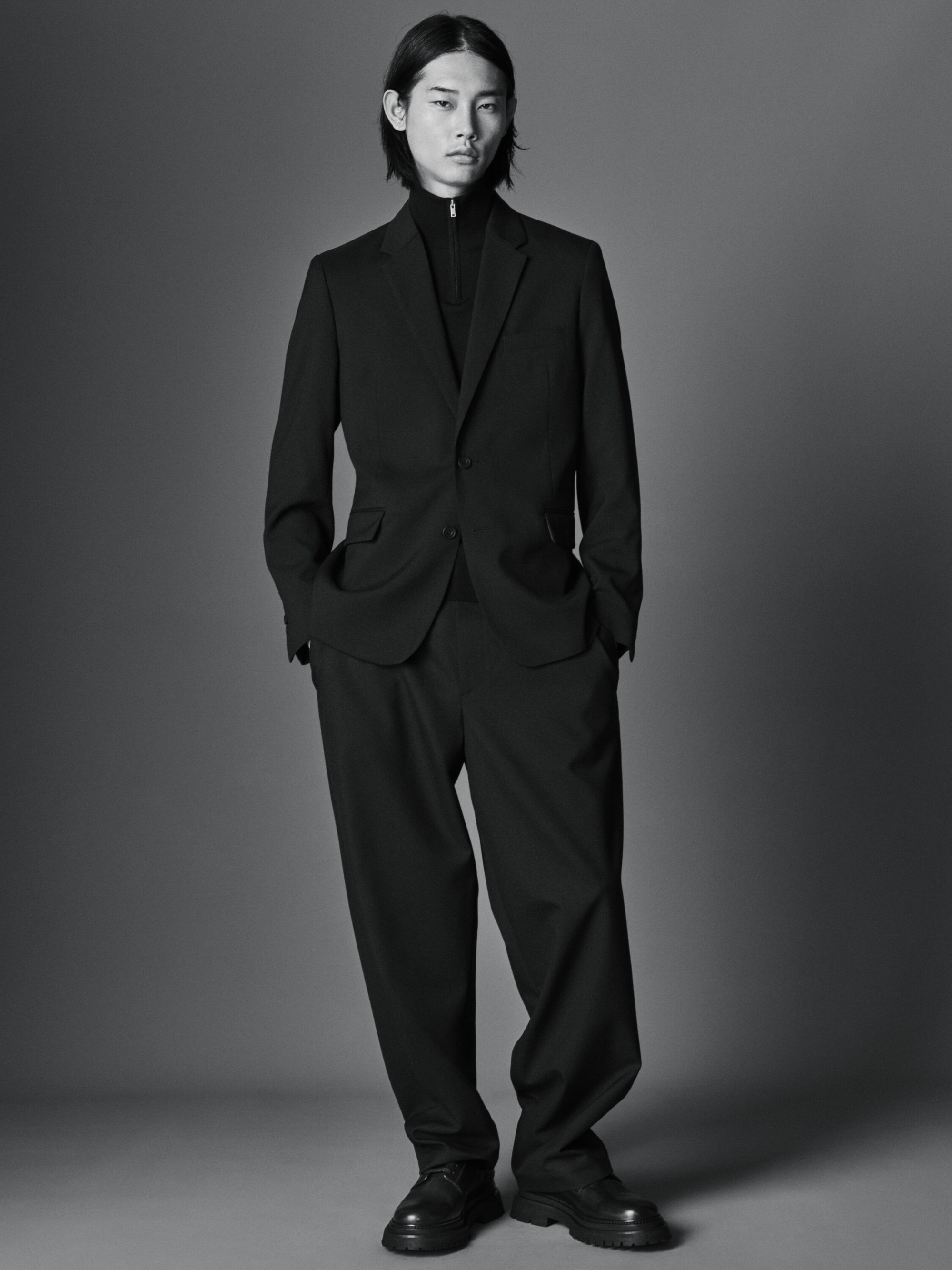 Man wearing a black suit and zip-up collar top against a plain studio background for the COS Fall 2023 campaign.