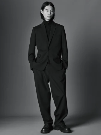 Man wearing a black suit and zip-up collar top against a plain studio background for the COS Fall 2023 campaign.