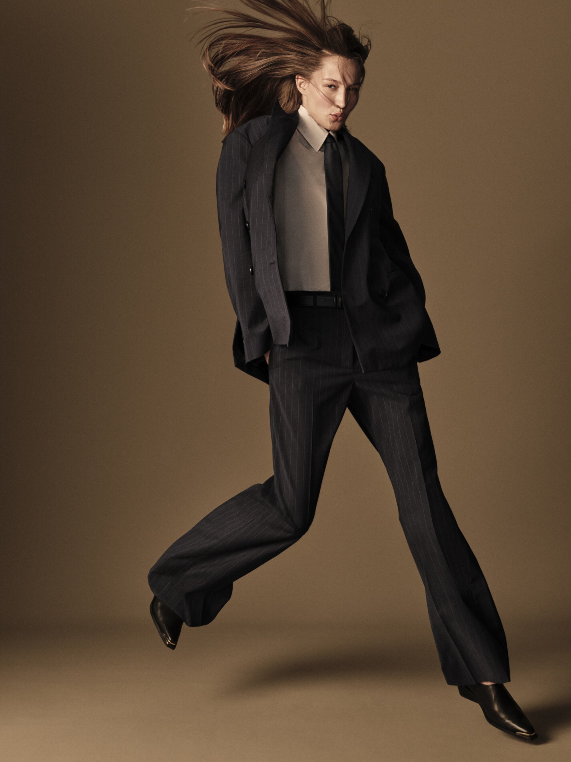 A slender, androgynous model leaps mid-air in a dark pinstripe suit, white shirt, and tie, against a neutral studio backdrop. Hair is windblown.