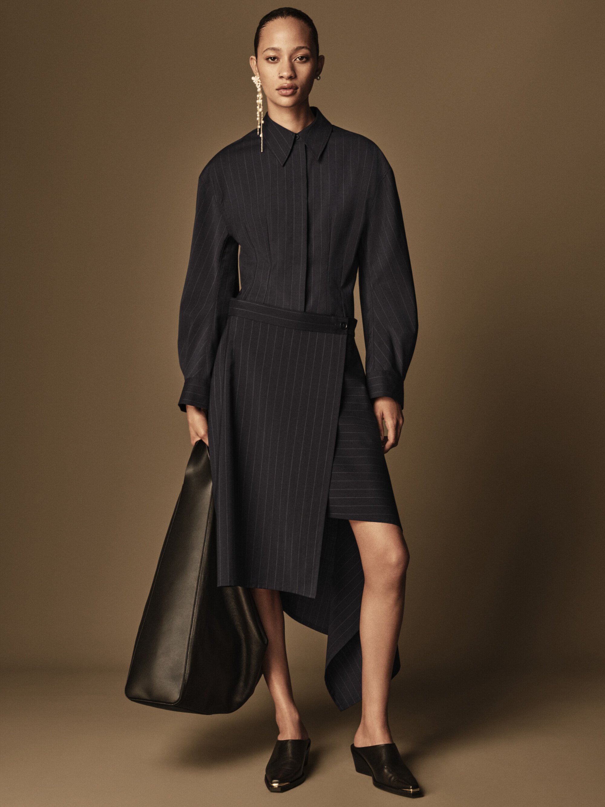 Fashion model wears a pinstripe dress, earring and black bag in a studio shot. The backdrop is a solid tan color in a full-length view.