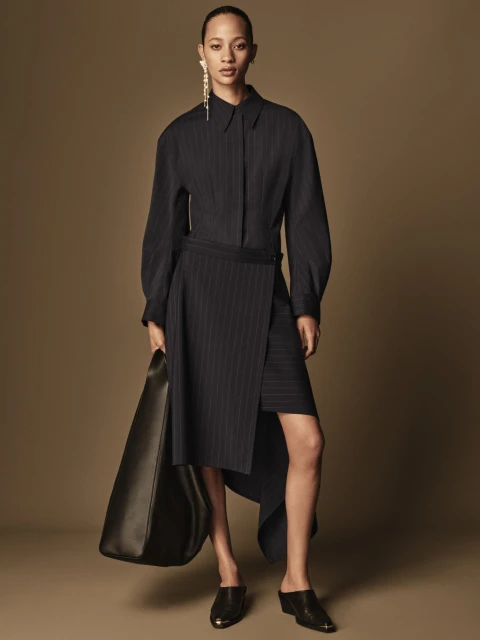 Fashion model wears a pinstripe dress, earring and black bag in a studio shot. The backdrop is a solid tan color in a full-length view.