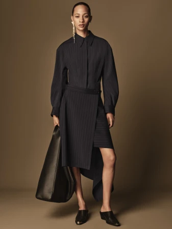 Fashion model wears a pinstripe dress, earring and black bag in a studio shot. The backdrop is a solid tan color in a full-length view.