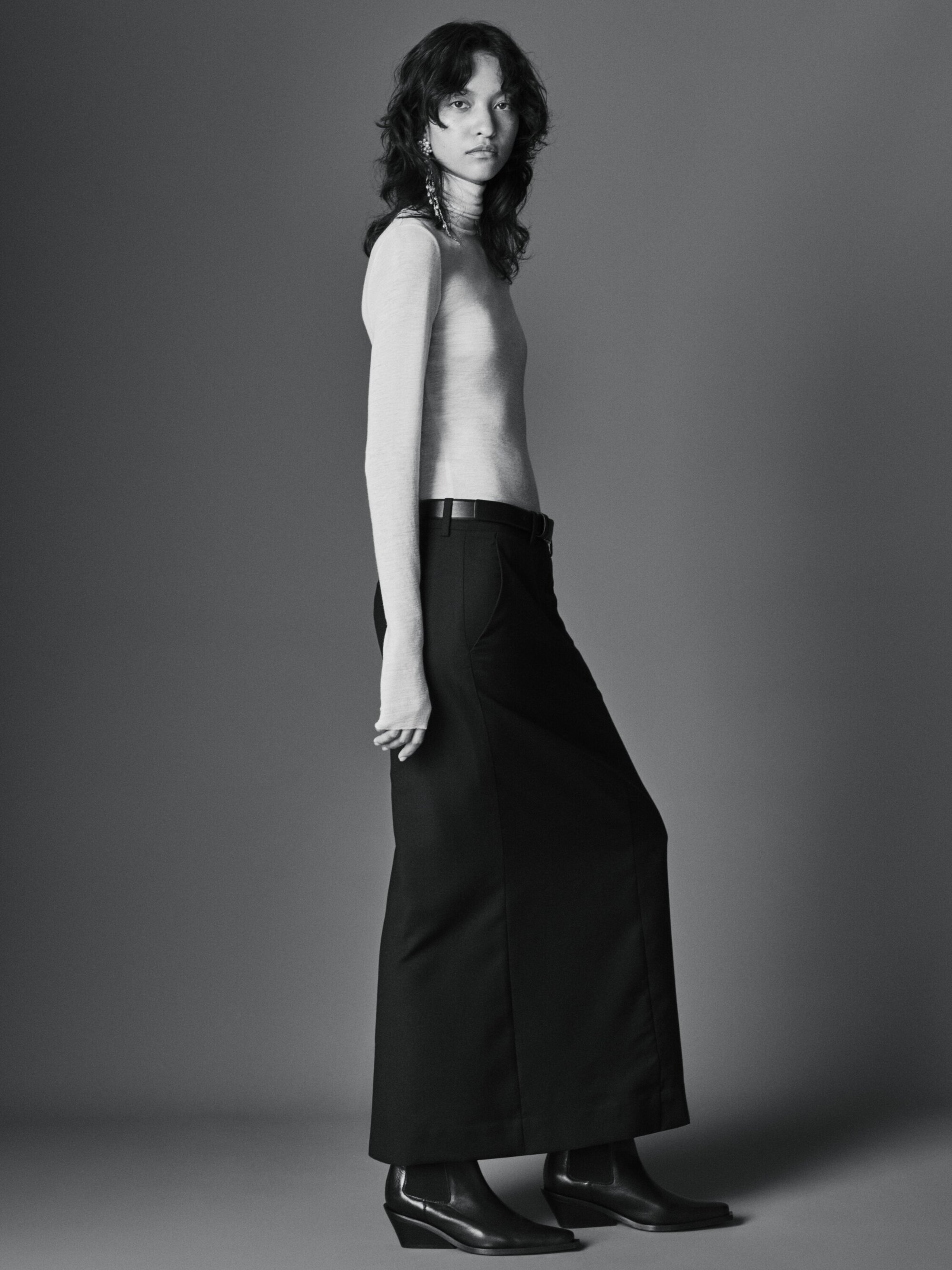 A solo female fashion model poses in a long black skirt and beige turtleneck in a studio with soft lighting and a neutral gray background.