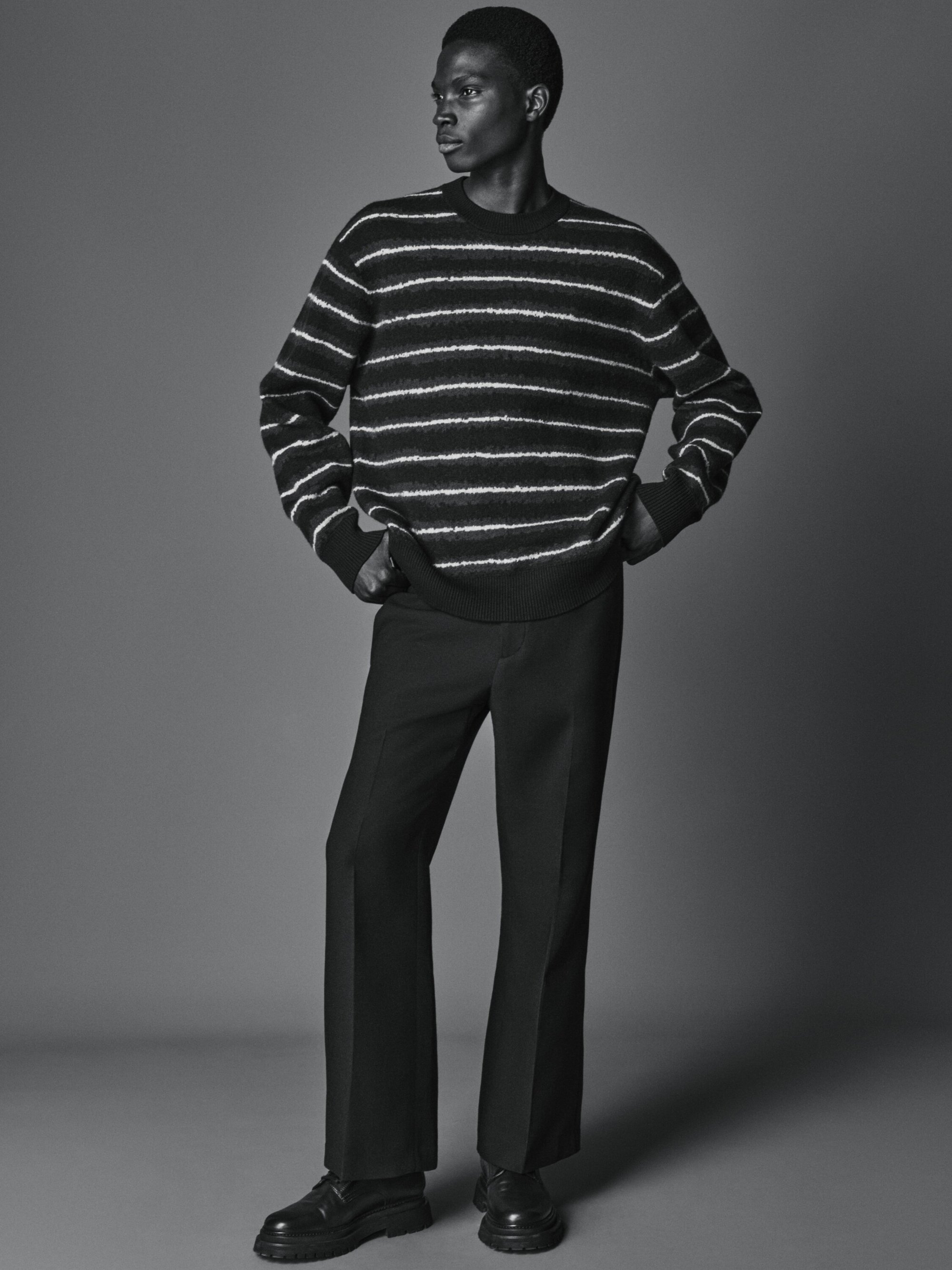 Full-length studio shot of a Black male model in a striped sweater and dress pants looking to his left, with a gray seamless background.