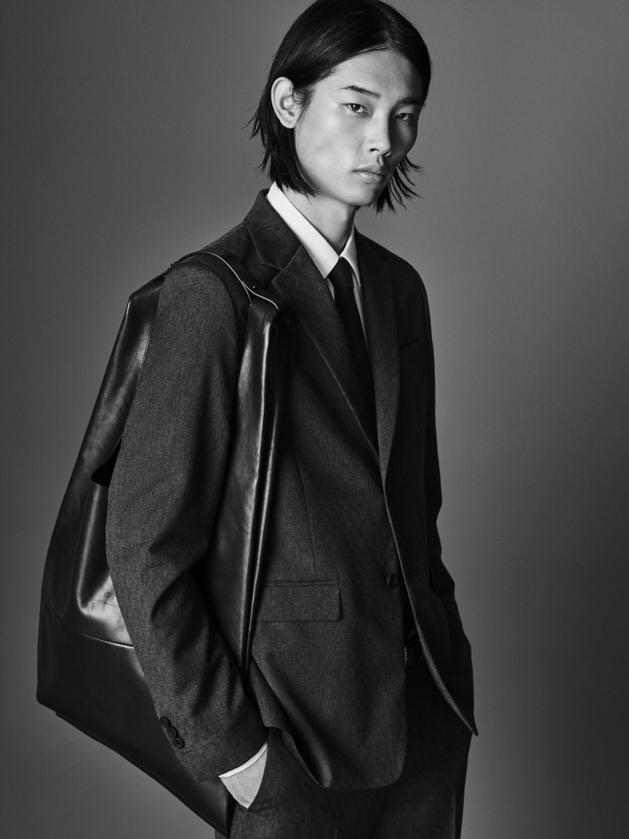 Portrait of a young adult male model in a dark suit with shoulder bag against a gray background, demonstrating fashion-forward elegance.