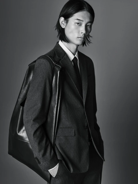 Portrait of a young adult male model in a dark suit with shoulder bag against a gray background, demonstrating fashion-forward elegance.