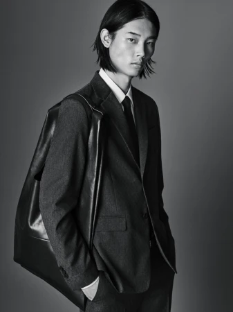 Portrait of a young adult male model in a dark suit with shoulder bag against a gray background, demonstrating fashion-forward elegance.