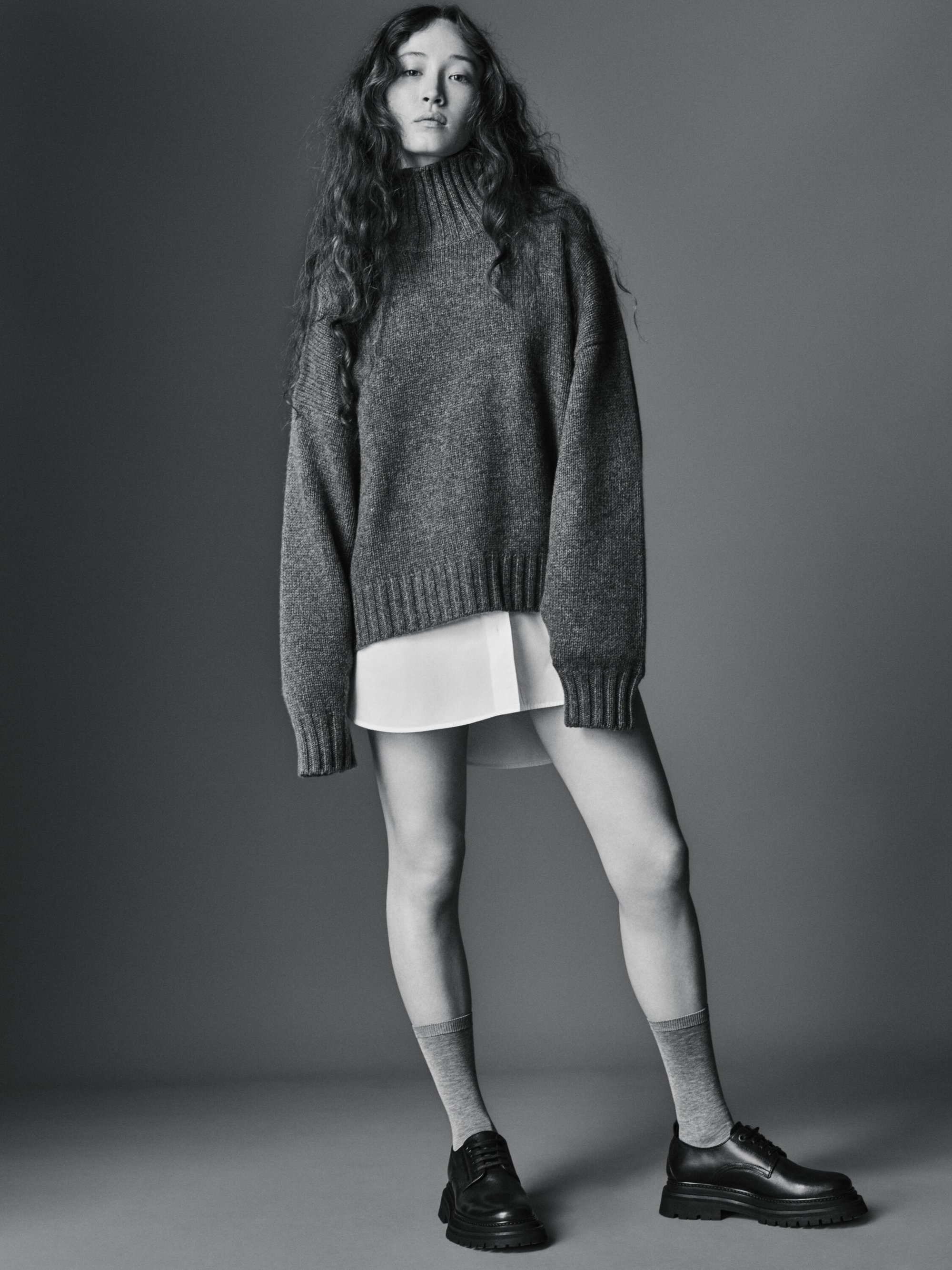 A young woman poses in a grey sweater, white shirt, grey socks, and black loafers against a grey background in a fashion studio setting.