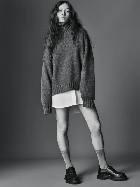 A young woman poses in a grey sweater, white shirt, grey socks, and black loafers against a grey background in a fashion studio setting.