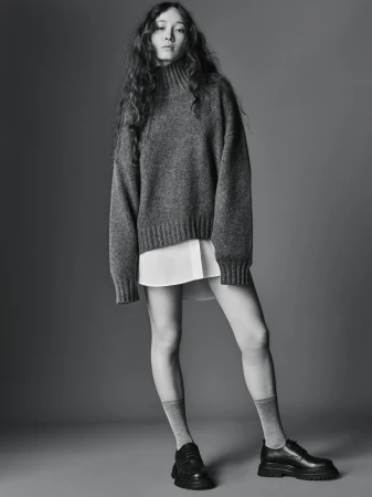 A young woman poses in a grey sweater, white shirt, grey socks, and black loafers against a grey background in a fashion studio setting.