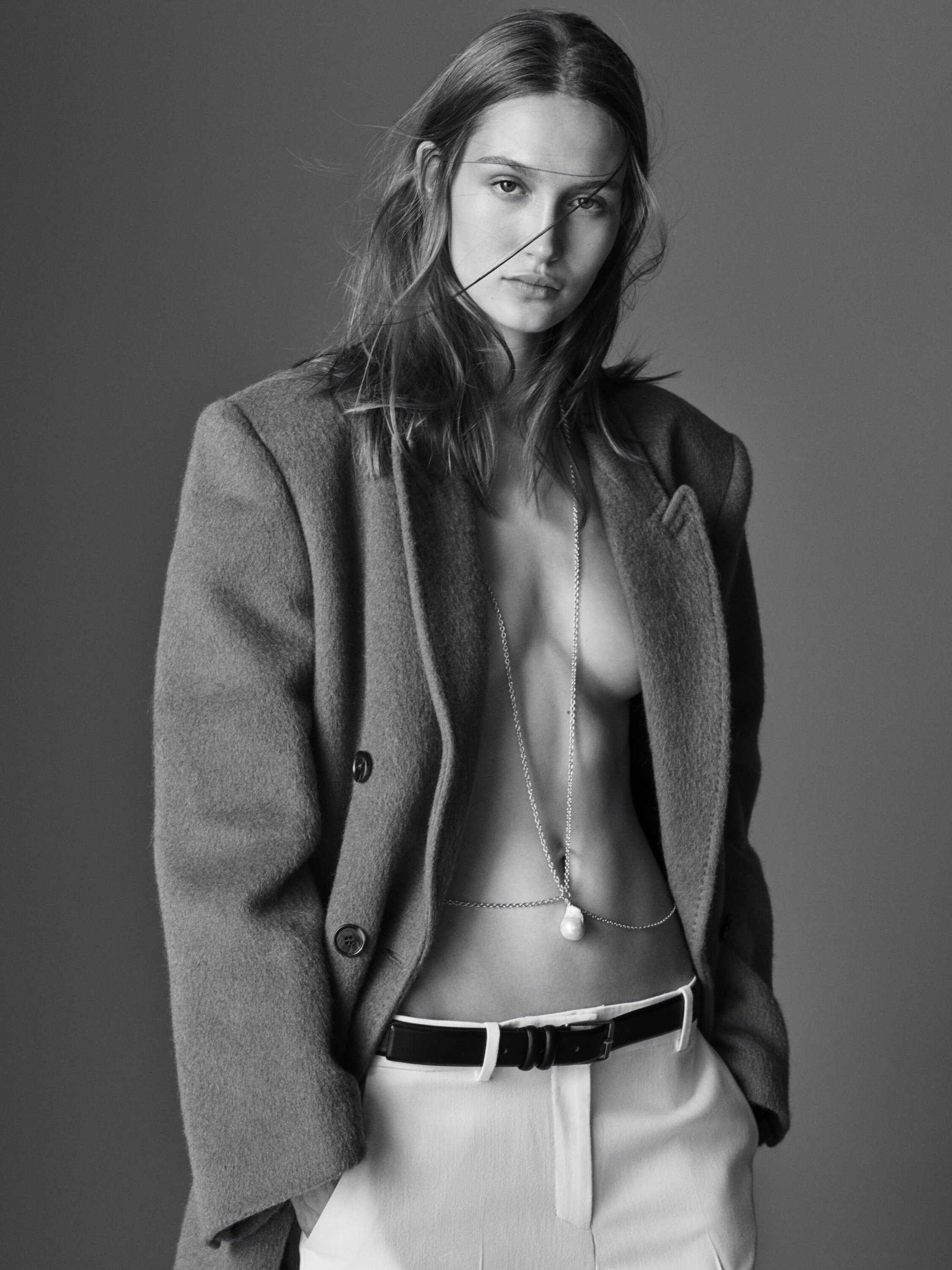 A fashion editorial shot showcases a Caucasian female model in an oversized blazer, white trousers, and a pearl necklace, against a grey backdrop.