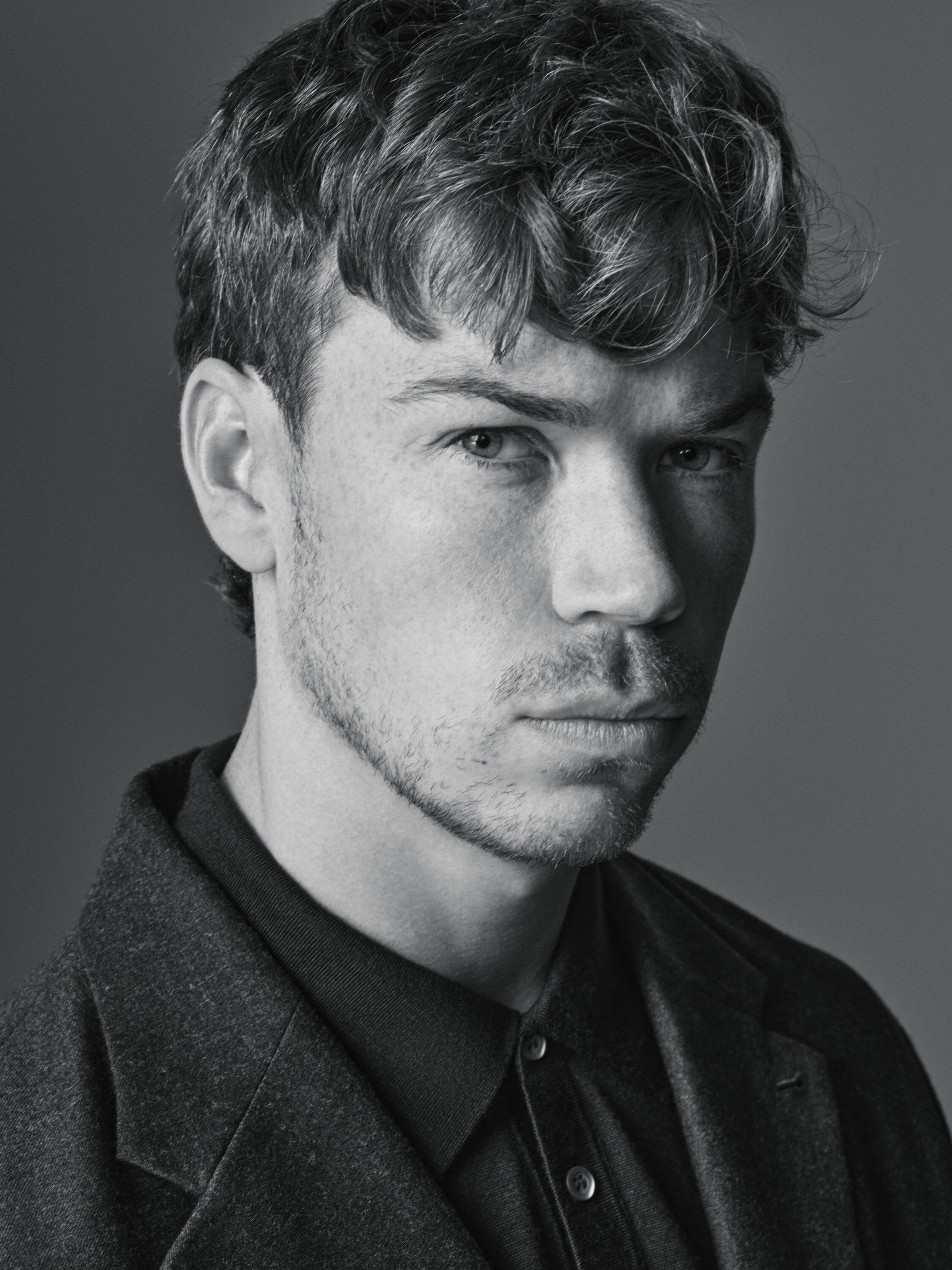 Monochrome portrait of a young adult man with short, wavy hair wearing a collared shirt and wool coat against a solid gray background.
