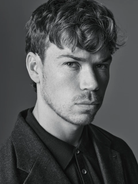 Monochrome portrait of a young adult man with short, wavy hair wearing a collared shirt and wool coat against a solid gray background.