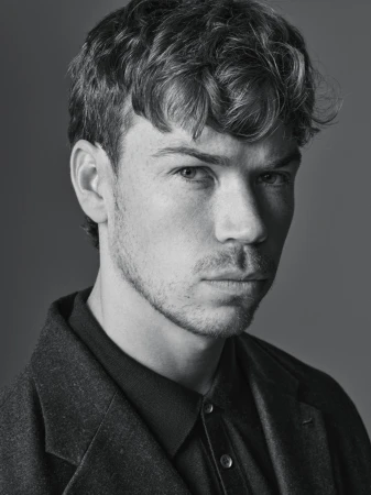 Monochrome portrait of a young adult man with short, wavy hair wearing a collared shirt and wool coat against a solid gray background.
