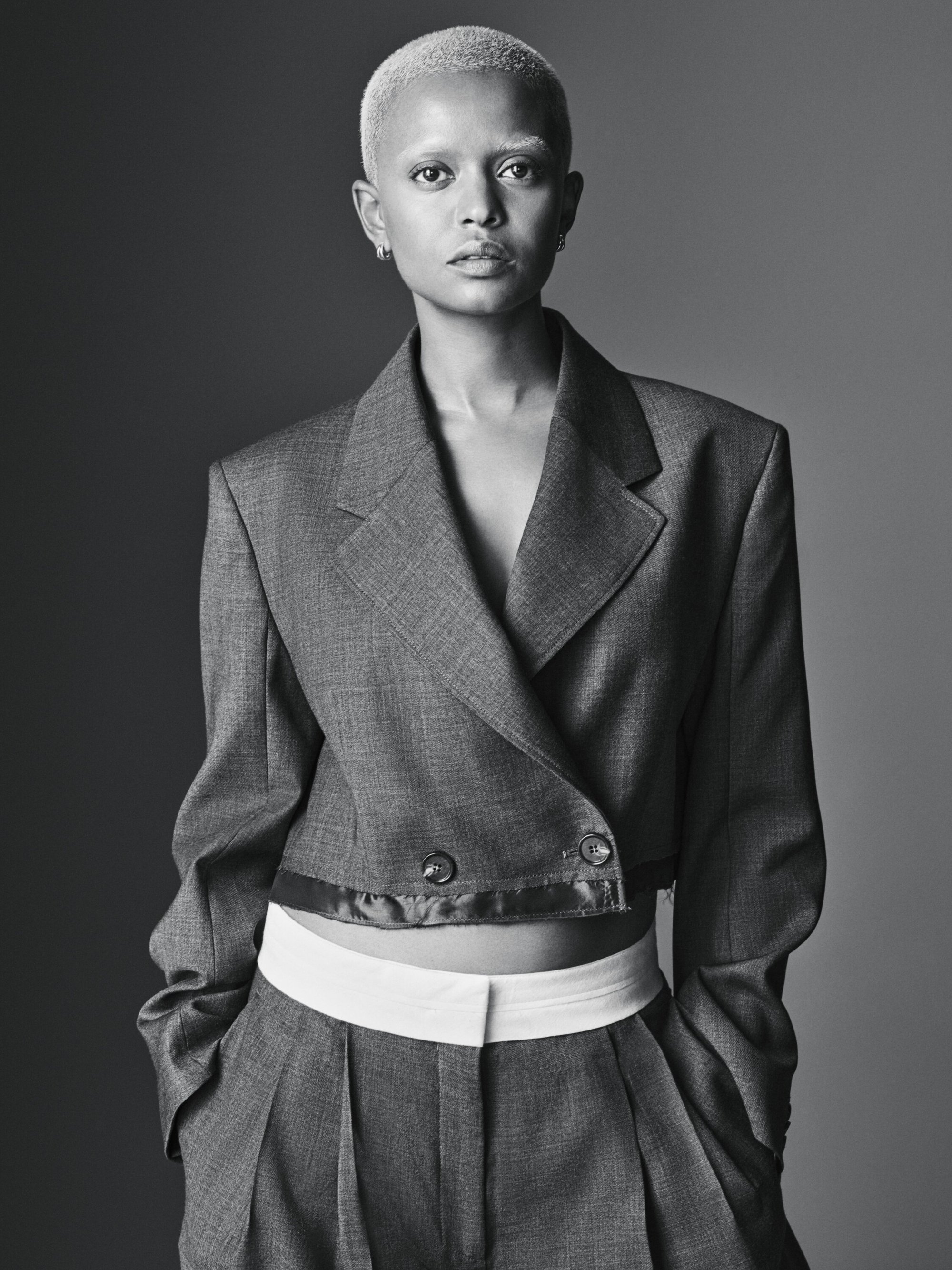A striking portrait of an adult female with a shaved head, wearing a gray suit with white undershirt against a solid gray background.
