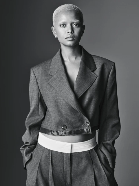 A striking portrait of an adult female with a shaved head, wearing a gray suit with white undershirt against a solid gray background.