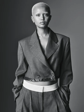 A striking portrait of an adult female with a shaved head, wearing a gray suit with white undershirt against a solid gray background.