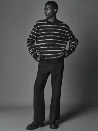 Full-length black-and-white portrait of a man standing in a striped knit sweater and tailored black trousers against a grey backdrop.
