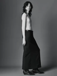 Model with dark wavy hair wearing a fitted long sleeve top and black maxi skirt against a solid grey background