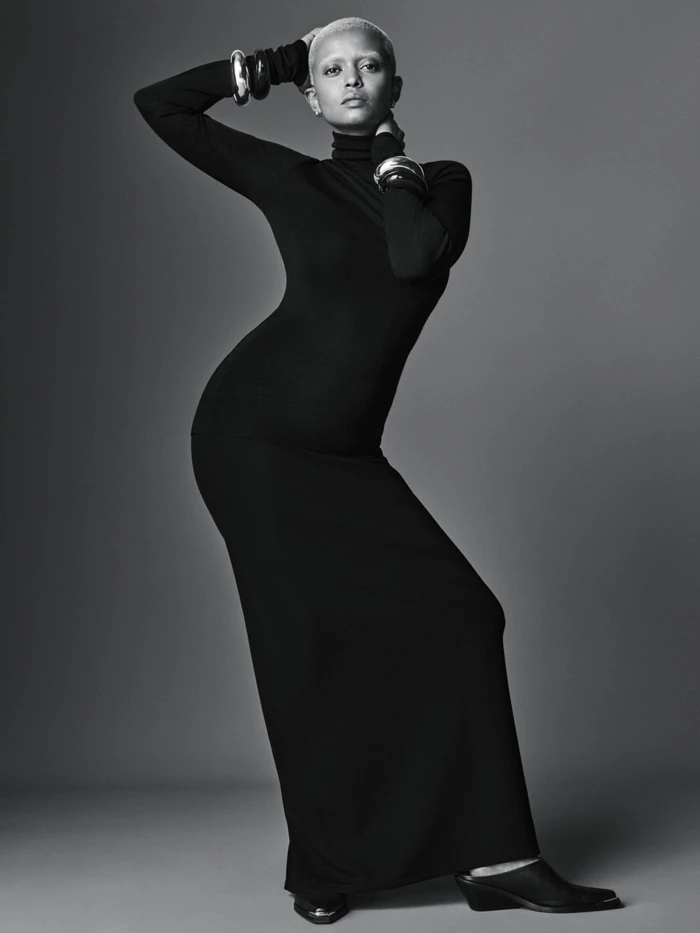 Model with short light hair wearing a black long-sleeve dress and silver bangles against a grey background.