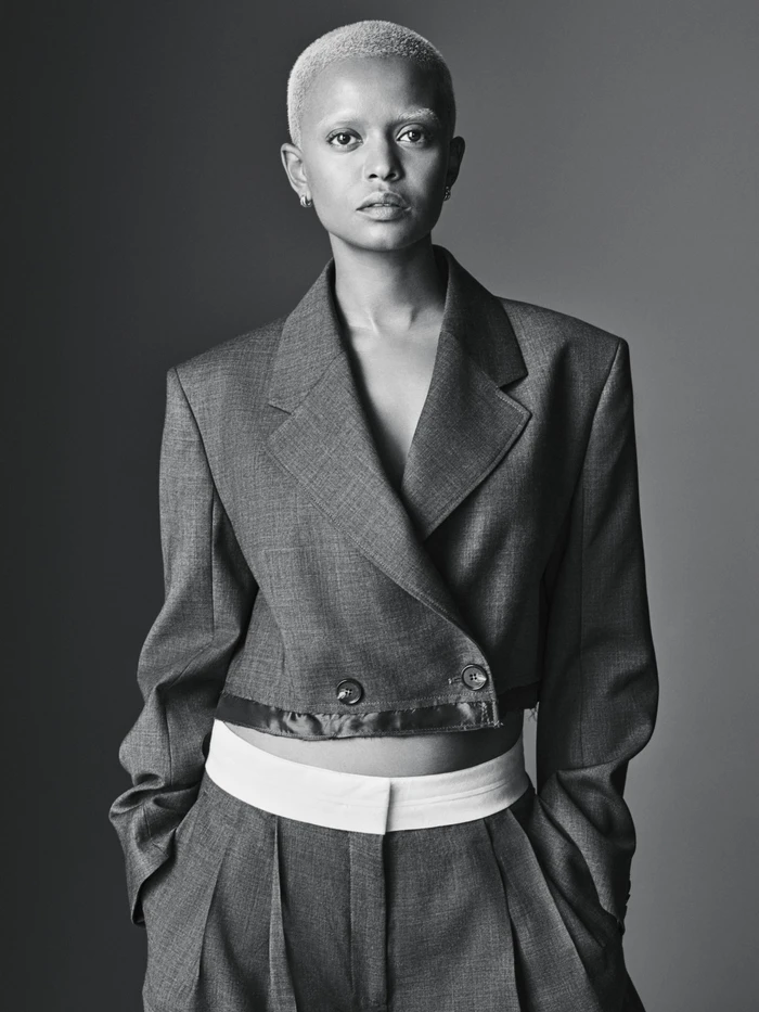 Kelela wearing a cropped tailored wool blazer and pleated trousers with hands in pockets against a studio backdrop.