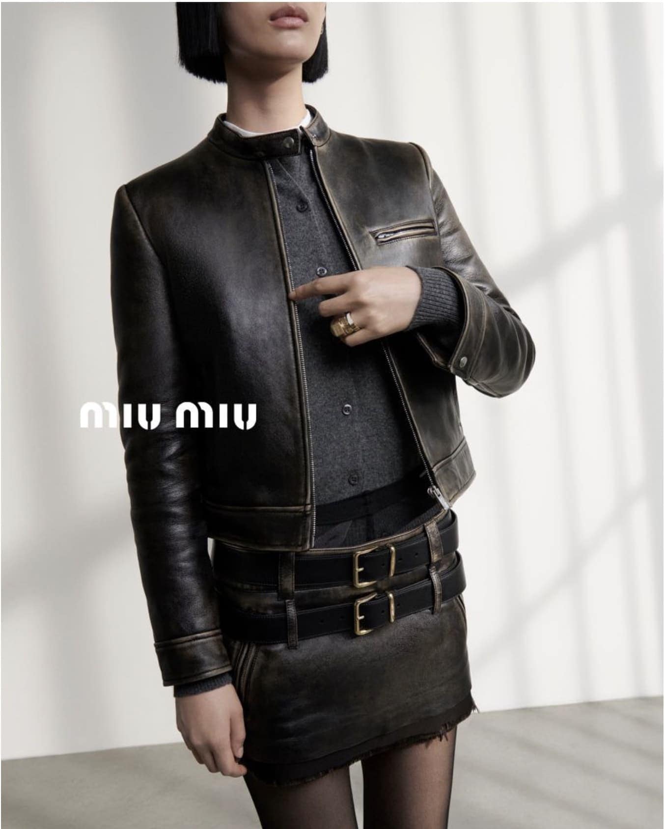 A woman wears a cropped leather jacket and a matching skirt with a double belt, a grey sweater, and black tights, standing indoors.