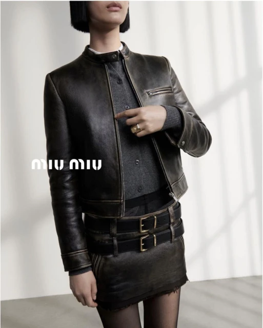A woman wears a cropped leather jacket and a matching skirt with a double belt, a grey sweater, and black tights, standing indoors.