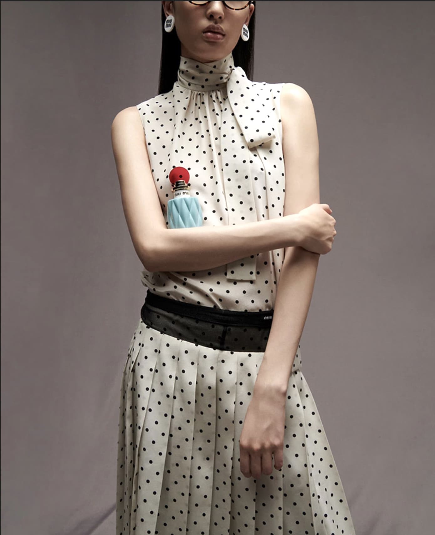 Woman in polka-dot dress holds perfume bottle in a studio shot, wearing glasses and earrings, with arms crossed.