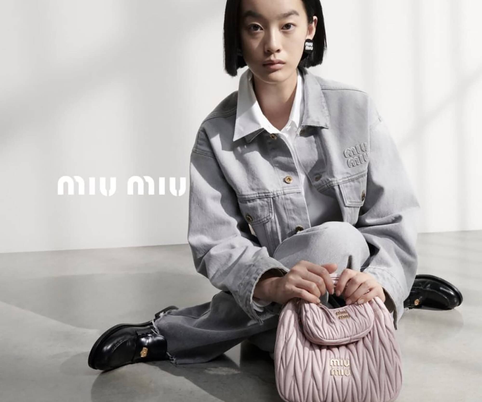 Fashion model poses indoors with a Miu Miu handbag, light grey jacket, and black shoes, sitting against a white wall, logo visible.