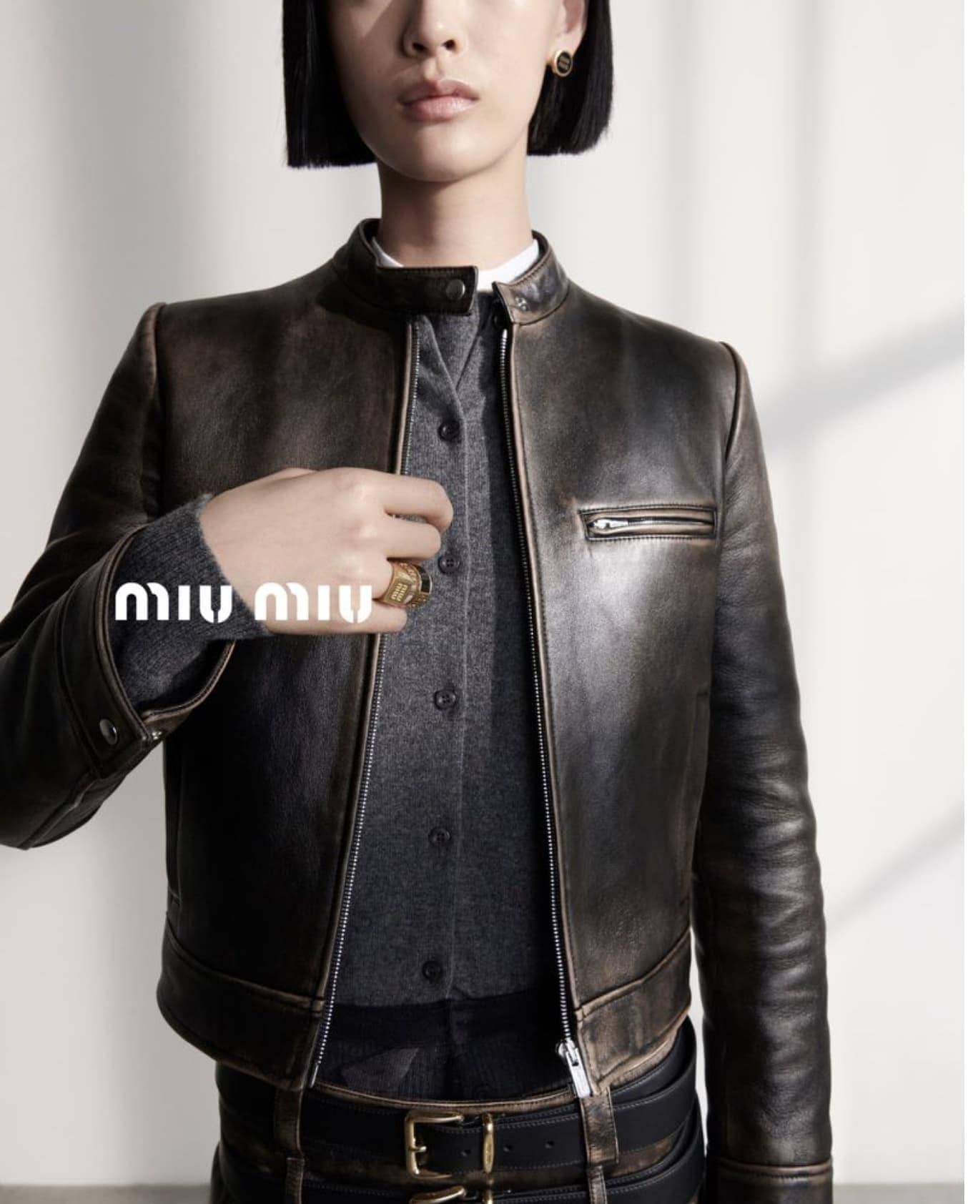 A model showcases a distressed-leather jacket with a gray button-down shirt, accessorized with a ring and earrings in a studio setting.