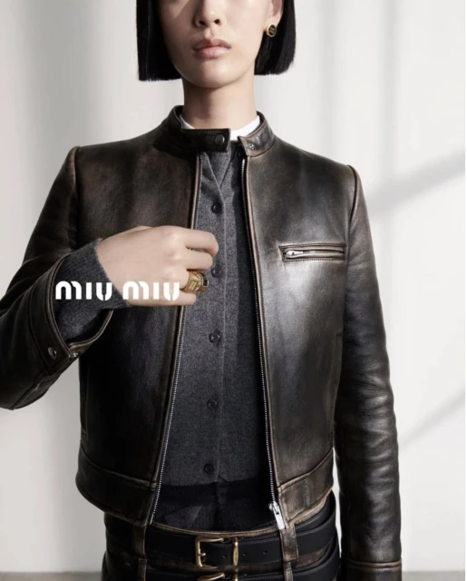 A model showcases a distressed-leather jacket with a gray button-down shirt, accessorized with a ring and earrings in a studio setting.