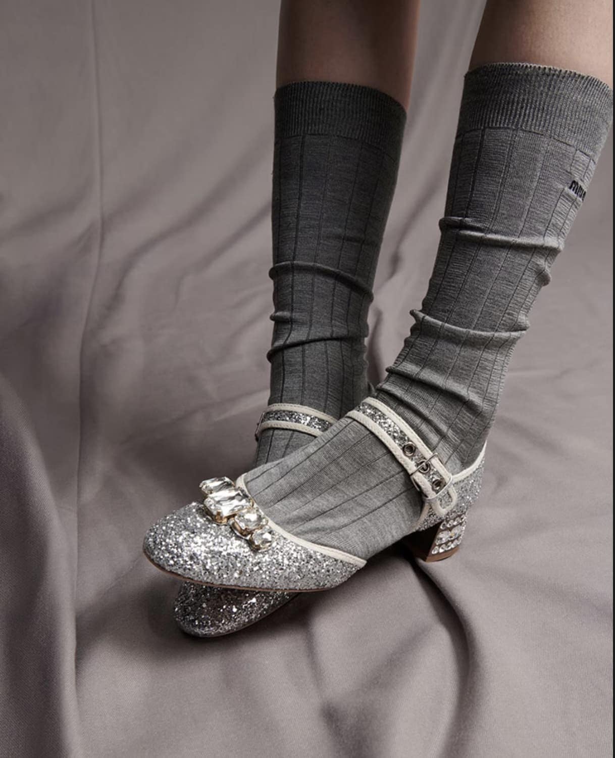 Close-up of sparkling silver Mary Jane heels, embellished with rhinestones, styled with tall grey socks against a grey fabric backdrop.