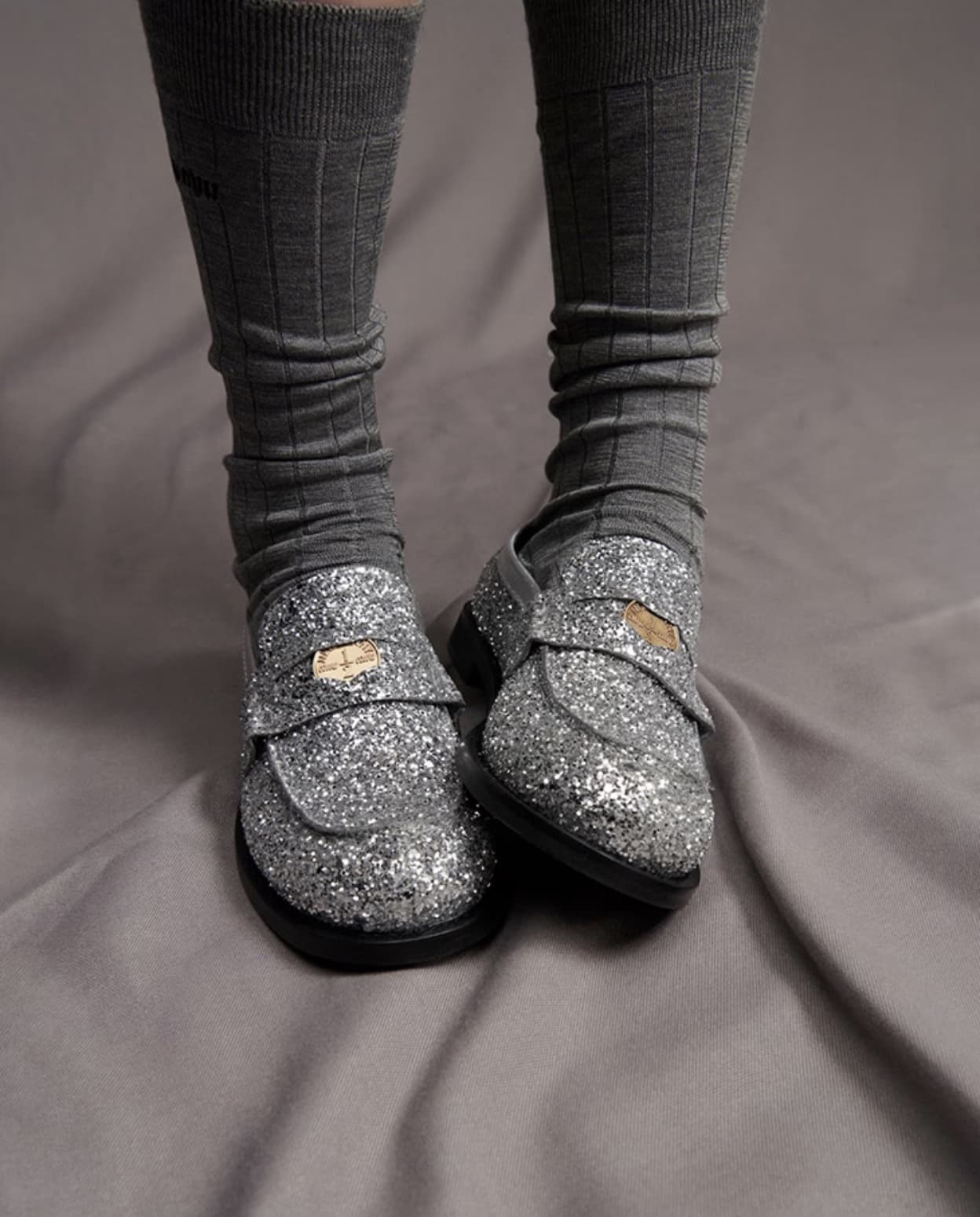 Close-up of a person's legs wearing gray socks and sparkling silver loafers on a gray wrinkled sheet, capturing a textured fashion detail.