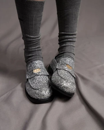 Close-up of a person's legs wearing gray socks and sparkling silver loafers on a gray wrinkled sheet, capturing a textured fashion detail.