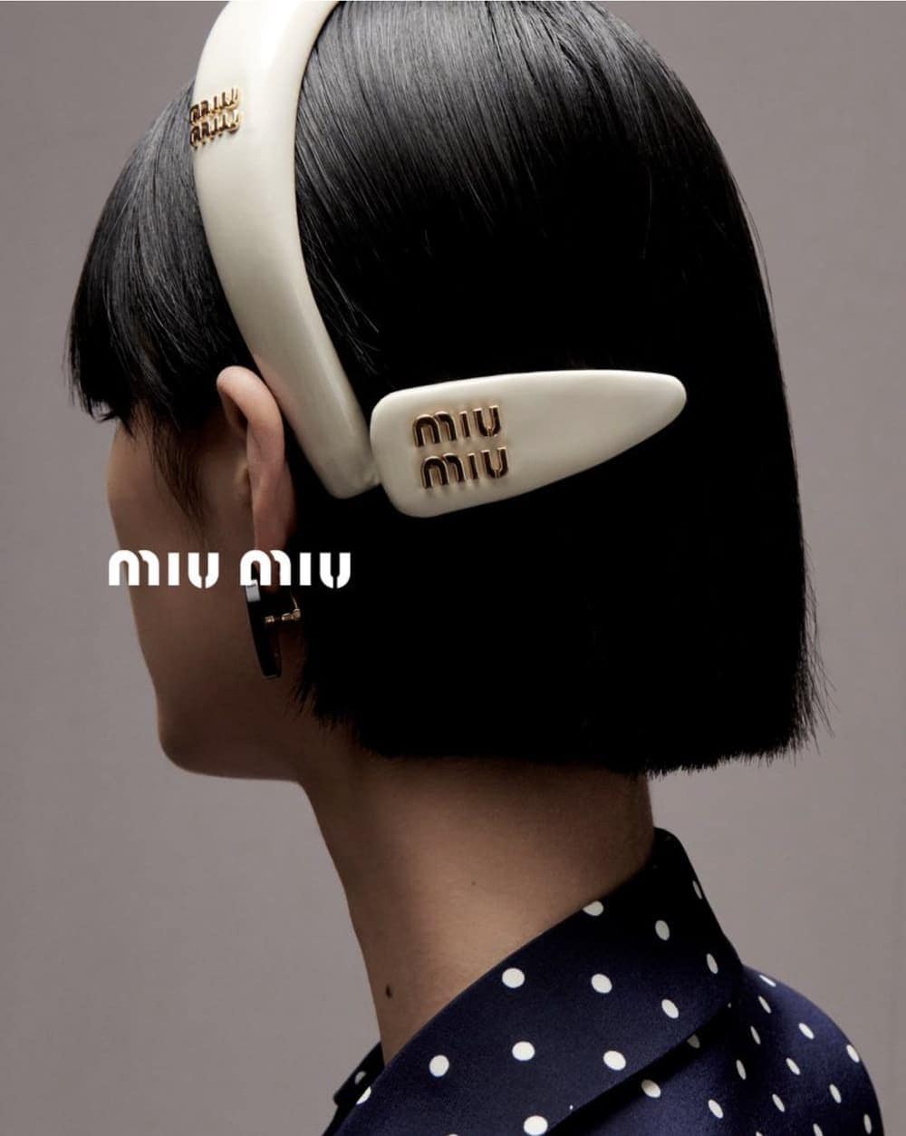 Woman with short black hair wearing a cream-colored Miu Miu headband and barrette with the brand's logo, against a gray backdrop.
