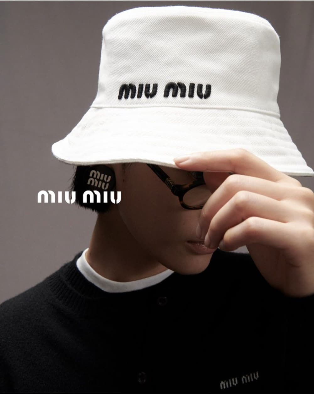 Person wearing a white Miu Miu bucket hat and black cardigan, posing against a grey backdrop in a close-up fashion shot.