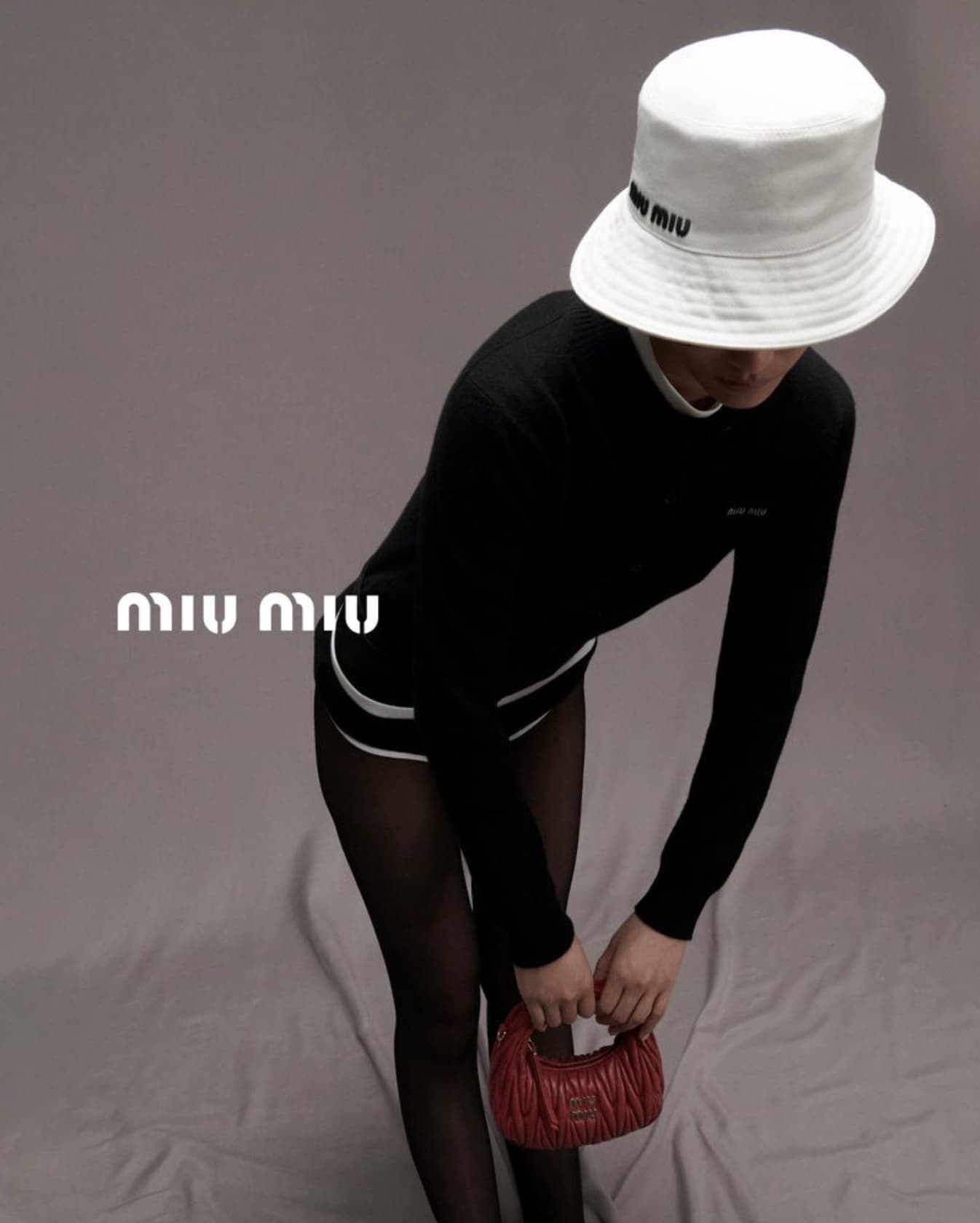 A slender person in black long-sleeve shirt, shorts, and white bucket hat holds a red leather purse against a light grey backdrop.