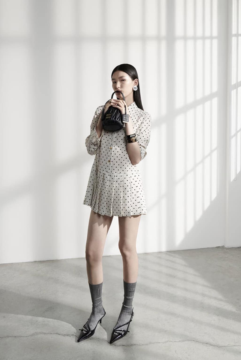 Full shot of a young Asian woman posing indoors, wearing a polka dot dress, grey socks, and black high heels while holding a small black purse.