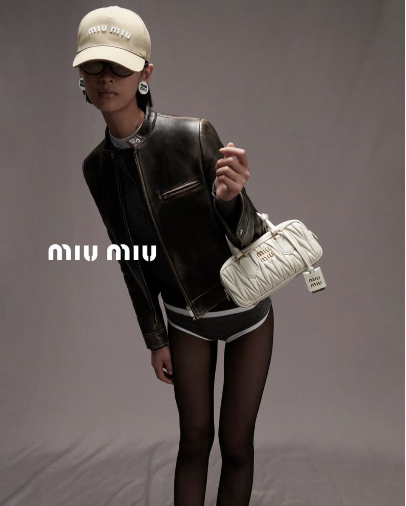 Woman in black jacket holds quilted Miu Miu bag, wearing a logo hat and dark tights, posing in a studio with a grey backdrop.