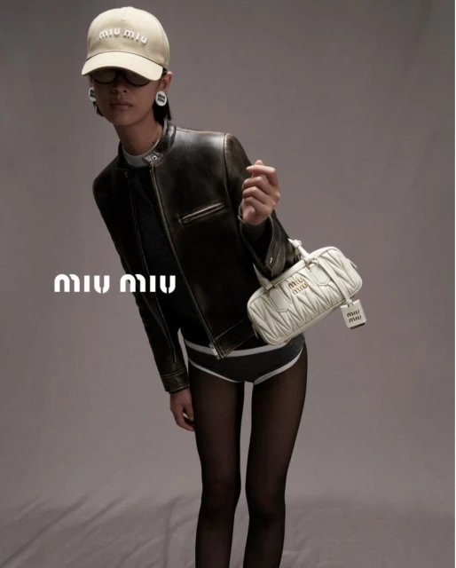 Woman in black jacket holds quilted Miu Miu bag, wearing a logo hat and dark tights, posing in a studio with a grey backdrop.