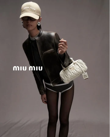 Woman in black jacket holds quilted Miu Miu bag, wearing a logo hat and dark tights, posing in a studio with a grey backdrop.