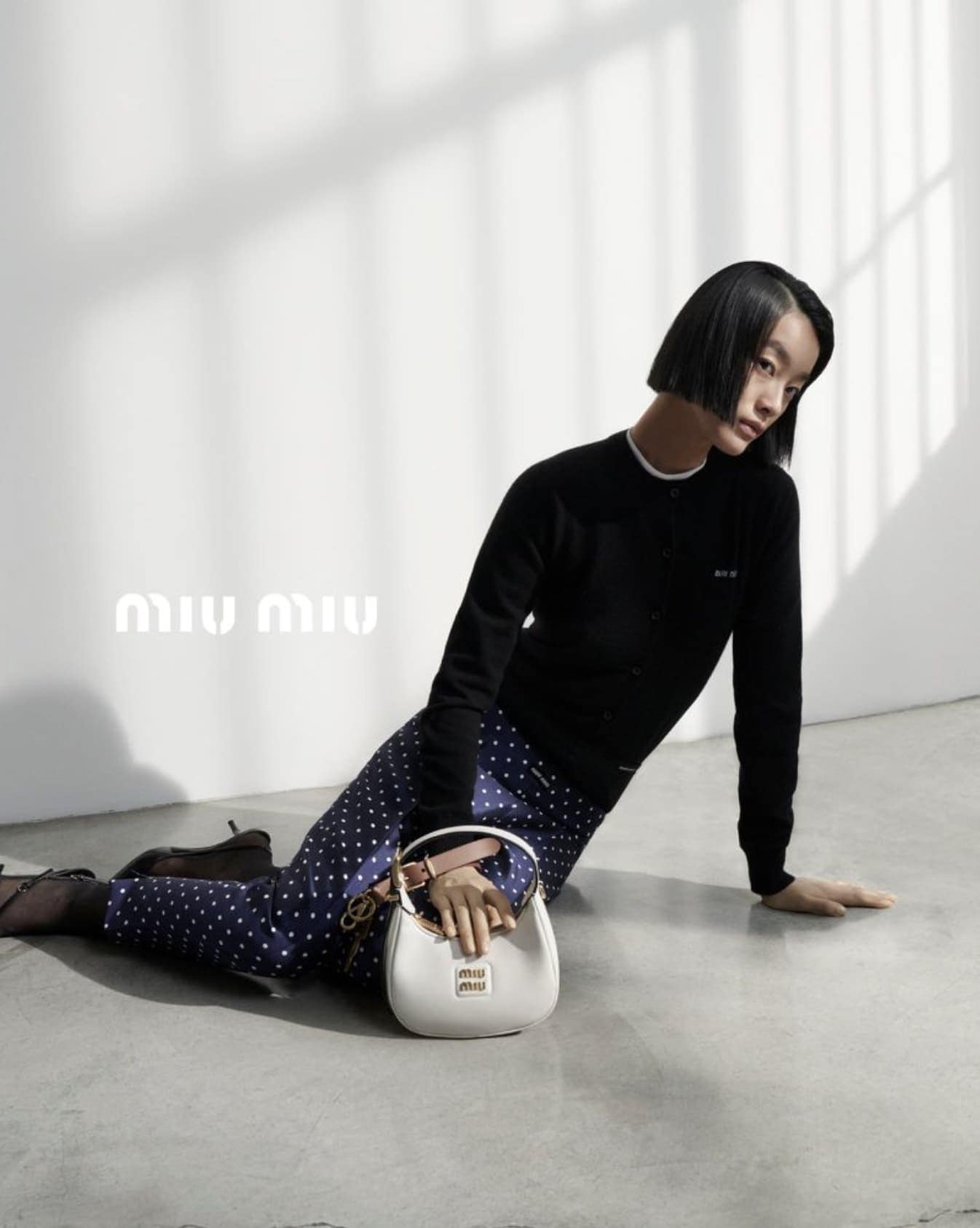 Fashion shot featuring an Asian female model lying on the floor with a white handbag, wearing a black cardigan and patterned pants in a studio setting.