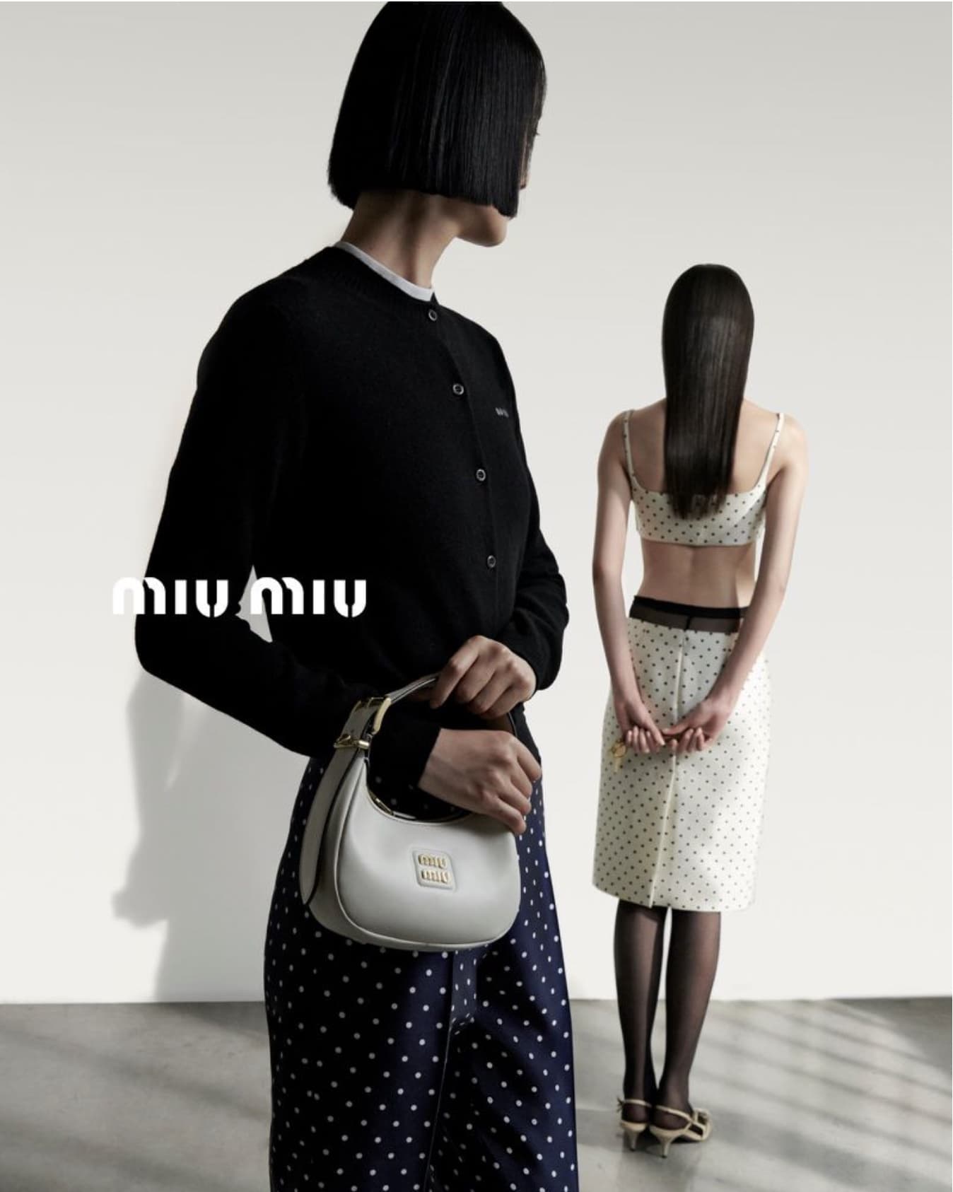 A Miu Miu fashion shot showcases two Asian women wearing black and white polka dot print skirt and cardigan with a white leather purse.